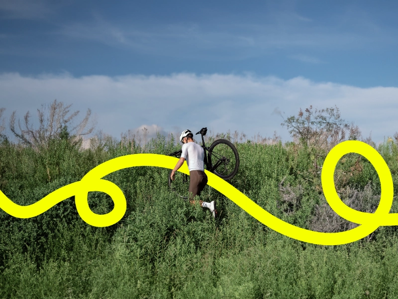 Radler trägt sein Gravelbike durchs Gestrüpp, eine gelbe JobRad Loop Linie kringelt sich durchs Bild Radler trägt sein Gravelbike durchs Gestrüpp, eine gelbe JobRad Loop Linie kringelt sich durchs Bild